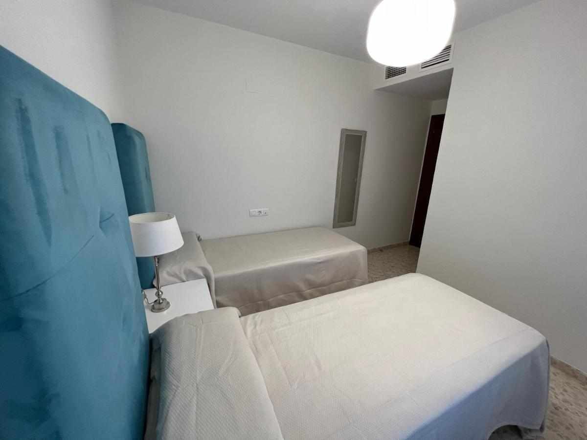 A small, simple room with two beds, a nightstand with a lamp, and a door leading to another room.