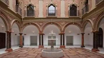 Un patio espacioso con una fuente central, rodeado de arcos y columnas, y un balcón en el nivel superior.