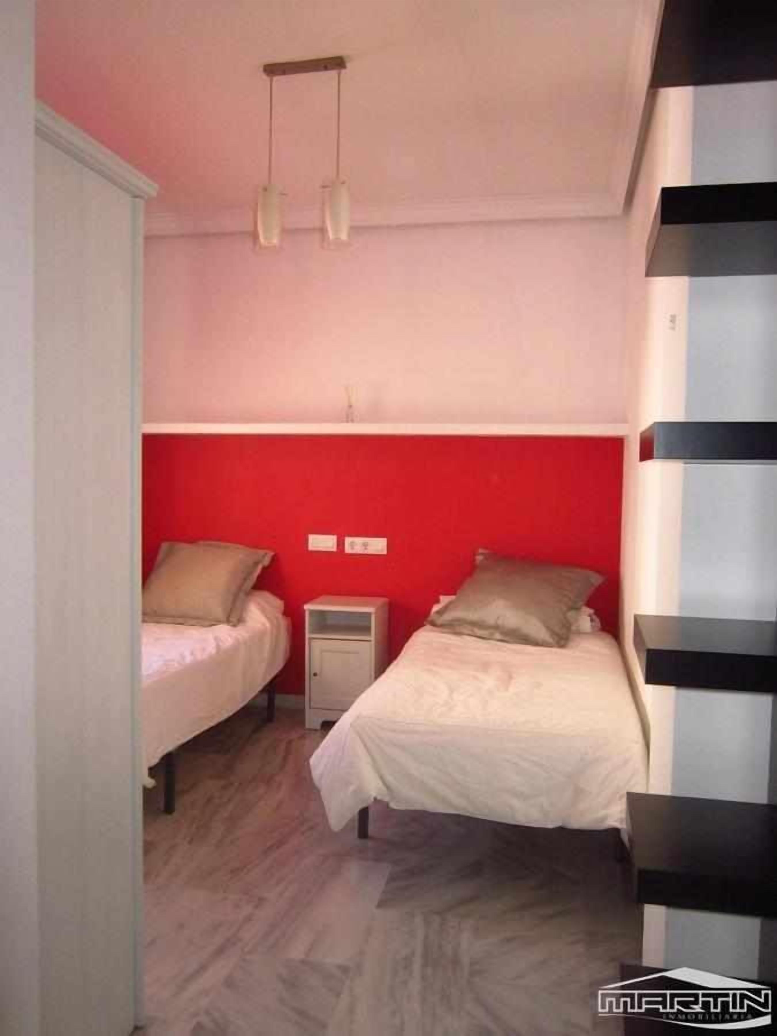 A small, compact room with two beds, a nightstand, and a red accent wall.