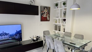 A modern living room with a glass dining table, grey chairs, a flat-screen TV, and a white shelving unit with decorative items. - APARTMENT QUINTANA CENTER (Jauja)