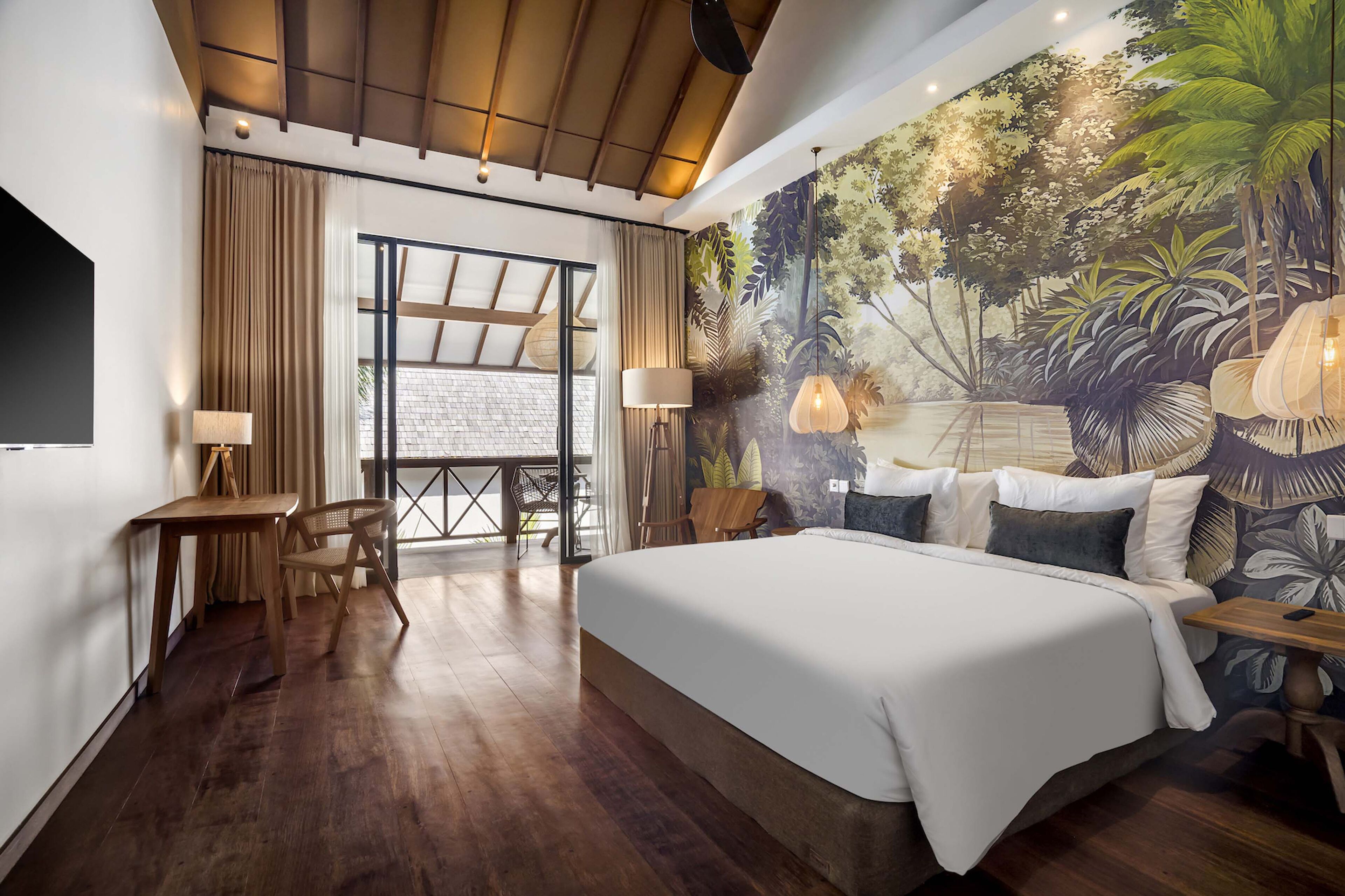 A bedroom with a large bed, wooden floor, a desk with a chair, a table, and a mural of a tropical scene.