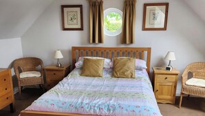Une chambre à coucher avec un lit en bois, deux tables de chevet, deux fauteuils en osier, une fenêtre avec des rideaux et des tableaux encadrés accrochés au mur.