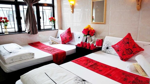 A hotel room with two beds, each with a red runner, white linens, and red floral pillows. There are two bedside tables with red cups and a vase of flowers. - Davao Hostel (Kowloon)