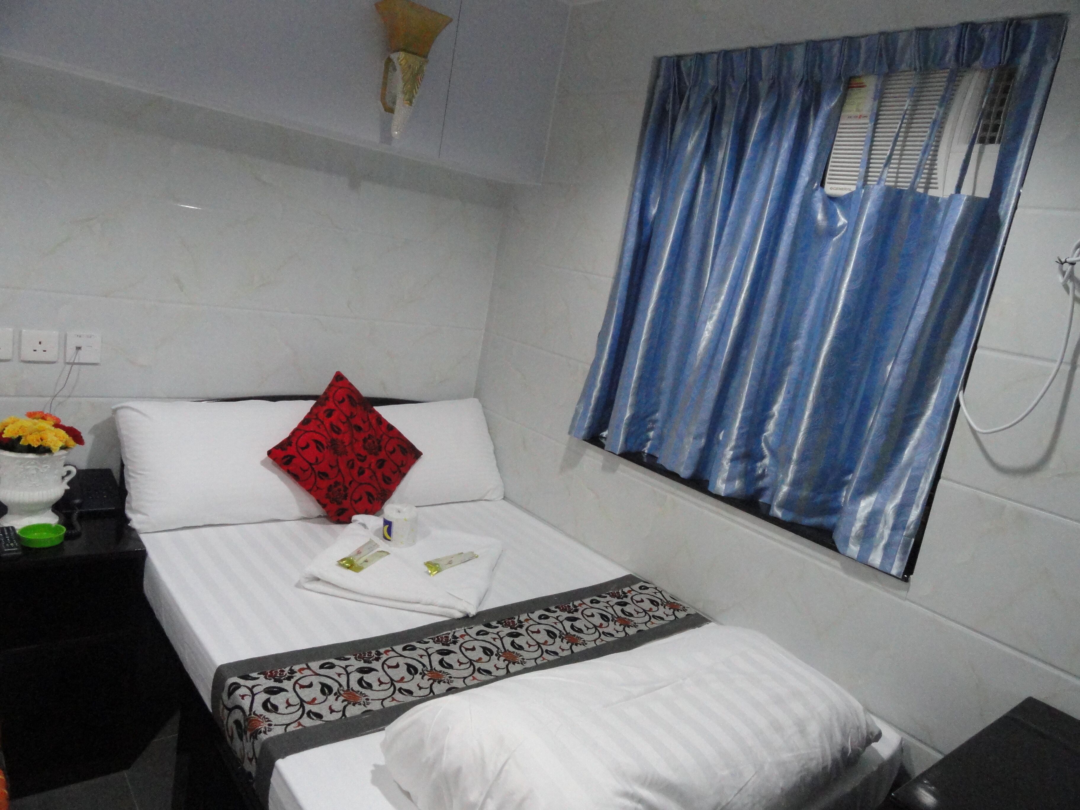 Une chambre d’hôtel avec un lit, une petite table sur laquelle est posé un vase de fleurs, un oreiller rouge et une fenêtre avec des rideaux bleus.