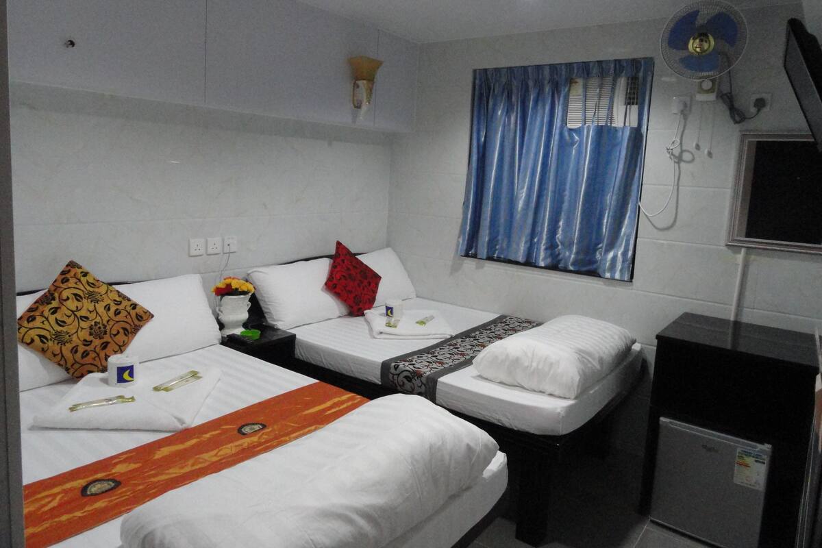 Habitación de hotel con dos camas individuales, una mesita con un florero y un ventilador de pared, además de una ventana con cortinas azules.