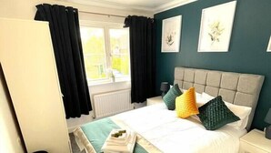 A neatly arranged bedroom with a bed, bedside tables, a radiator, and a window with curtains. - Remarkable 5-Bed House in Horley (Horley)