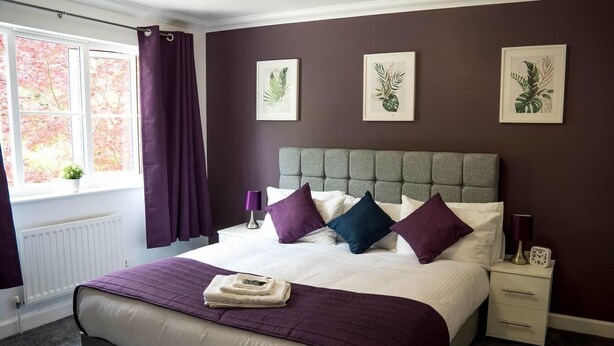 A neatly made bed with a grey headboard, white bedding, and purple accents. There are three framed botanical prints on the wall, a window with purple curtains, and a bedside table with a lamp and a clock. - Remarkable 5-Bed House in Horley (Horley)