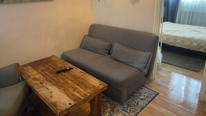 Une pièce chaleureuse avec un canapé gris, une table en bois sur laquelle se trouve une télécommande, un tapis à motifs et un lit recouvert d’une couvre-lit à fleurs.