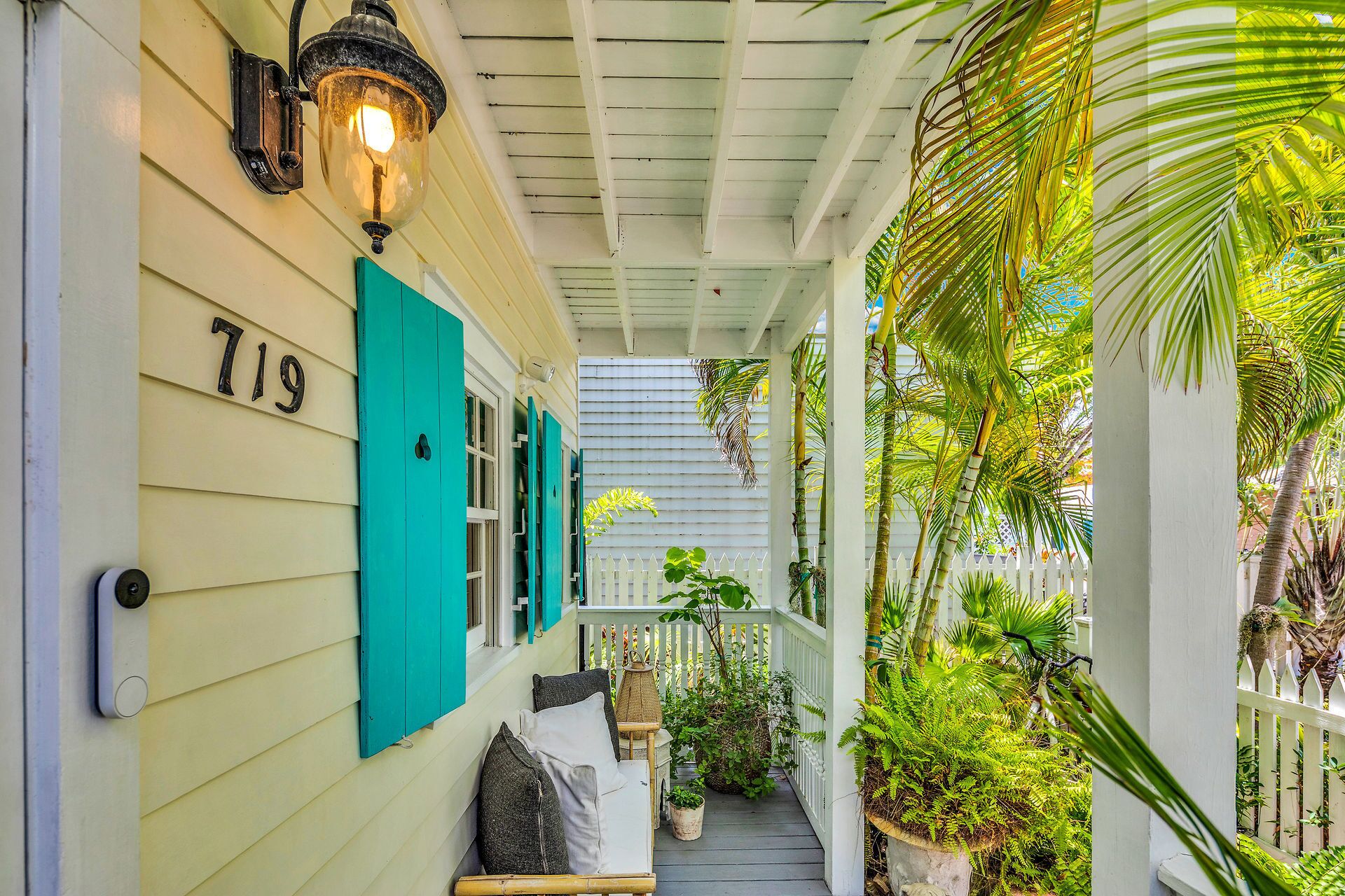 A porch with teal shutters, a white picket fence, and lush tropical plants.