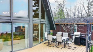 A modern outdoor deck with a dining area, including chairs and a table, and a view of the interior through large glass doors. - Caribou Lake A-Frame w/ Sauna, Heirloom Vacations (Duluth)