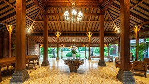 A covered outdoor area with wooden pillars, a central table with a flower vase, and benches. - Griya Shanti Suites (Denpasar)