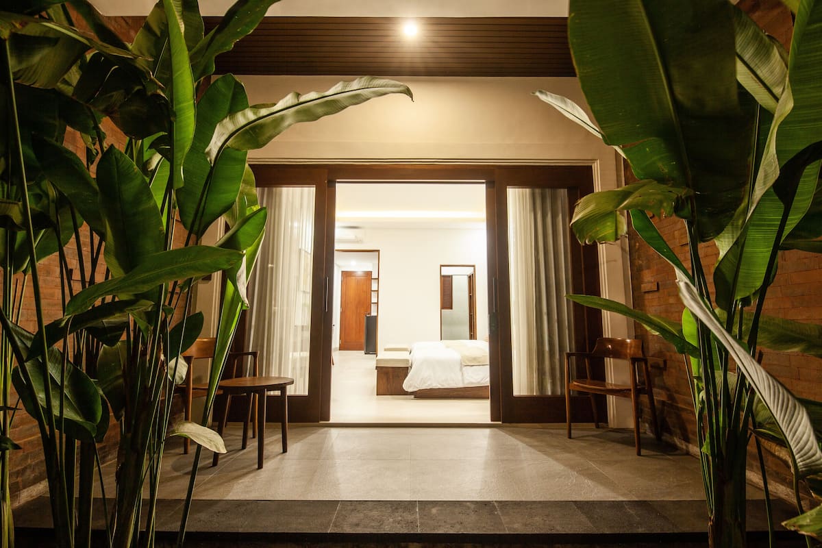 A hotel room with a bed, a desk, and chairs. There are large green plants in the foreground.