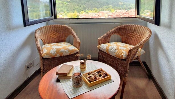 A room with two wicker chairs with floral cushions, a small round table with a book, a jar, and a wooden tray with snacks. - La Rectoral Arriondas 2 (Arriondas)