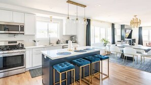 A modern kitchen with white cabinets, a central island with bar stools, a dining area with a table and chairs, and a large window providing natural light.