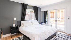 A bedroom with a bed, a nightstand, a window with curtains, and a view of the outside.