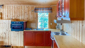 A compact kitchen with wooden walls, a built-in oven, a red cabinet, a window with a plaid curtain, and a stove.
