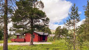 A red cabin with a porch, surrounded by trees and a grassy area.