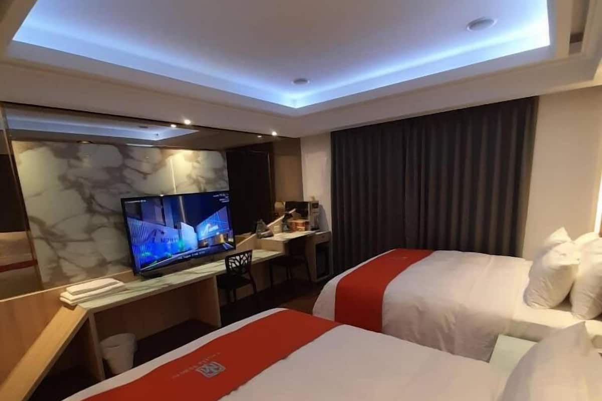 Une chambre d’hôtel avec deux lits, un bureau équipé d’un ordinateur et d’une télévision, et un plafond doté d’un éclairage encastré.