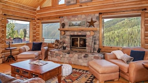 L’intérieur d’une cabane en rondins est agrémenté d’une cheminée en pierre, de canapés en cuir et de grandes fenêtres offrant une vue sur les montagnes.