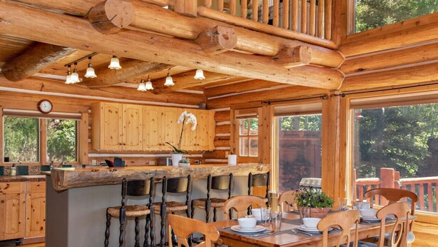 L’intérieur d’un chalet rustique est agrémenté d’une table à manger en bois, de tabourets de bar et d’une grande fenêtre donnant sur une zone boisée.