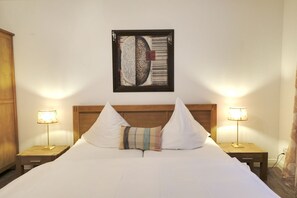 Ein ordentlich bezogenes Bett mit weißer Bettwäsche, zwei Nachttischlampen, einem Holz-Kopfteil und einem gerahmten Kunstwerk darüber.
