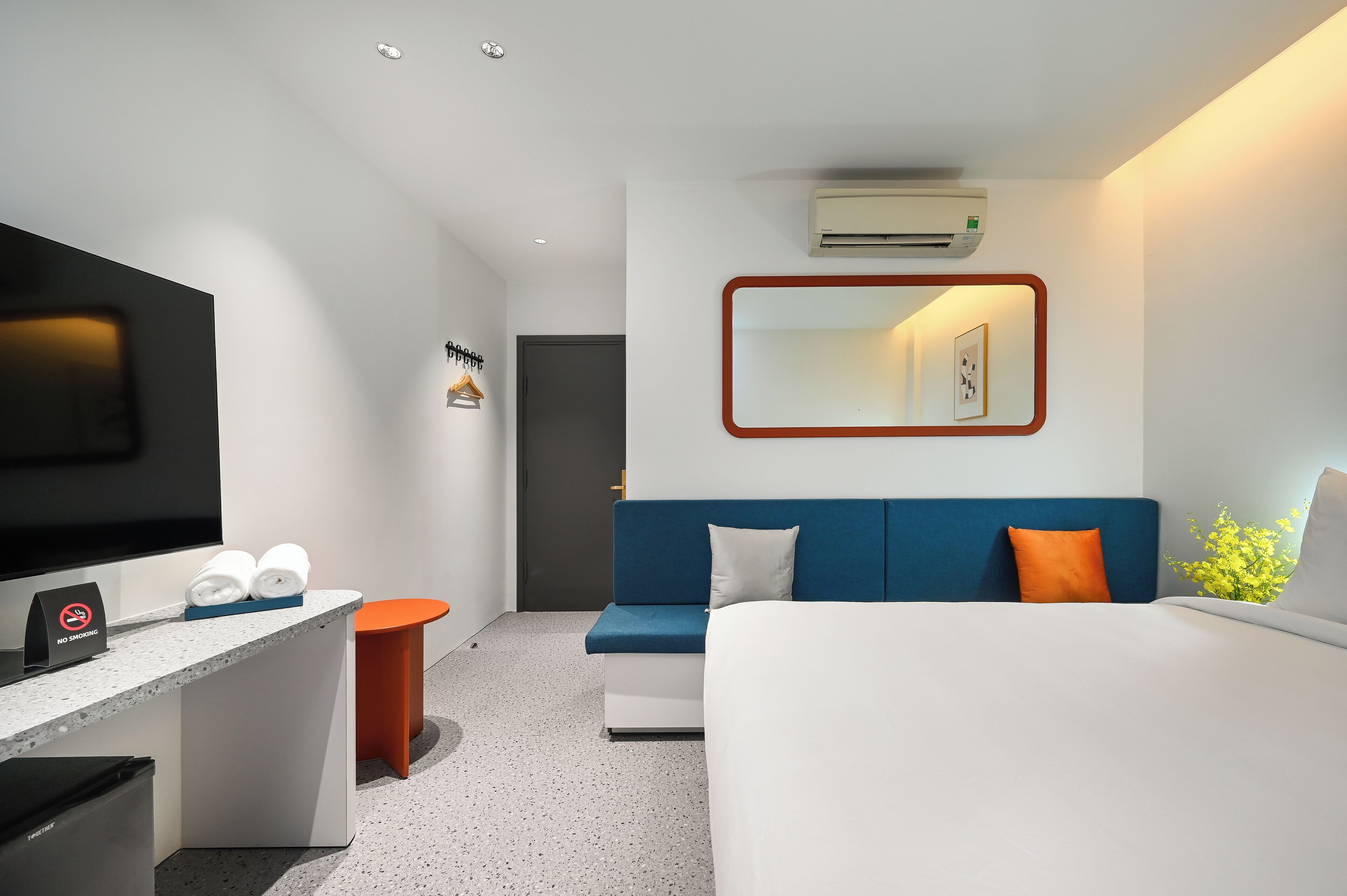 A modern hotel room with a flat-screen TV, a desk with a no smoking sign, a blue sofa, an orange stool, and a bed with white bedding.