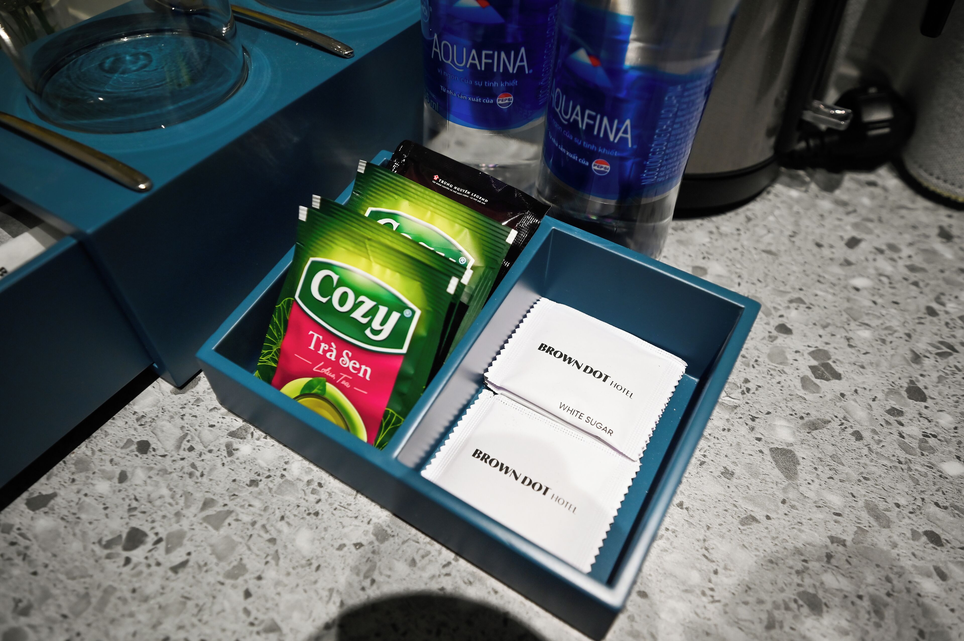 A blue box containing a Cozi tea bag, a Brown Dot Hotel white sugar packet, and a Brown Dot Hotel tea bag.