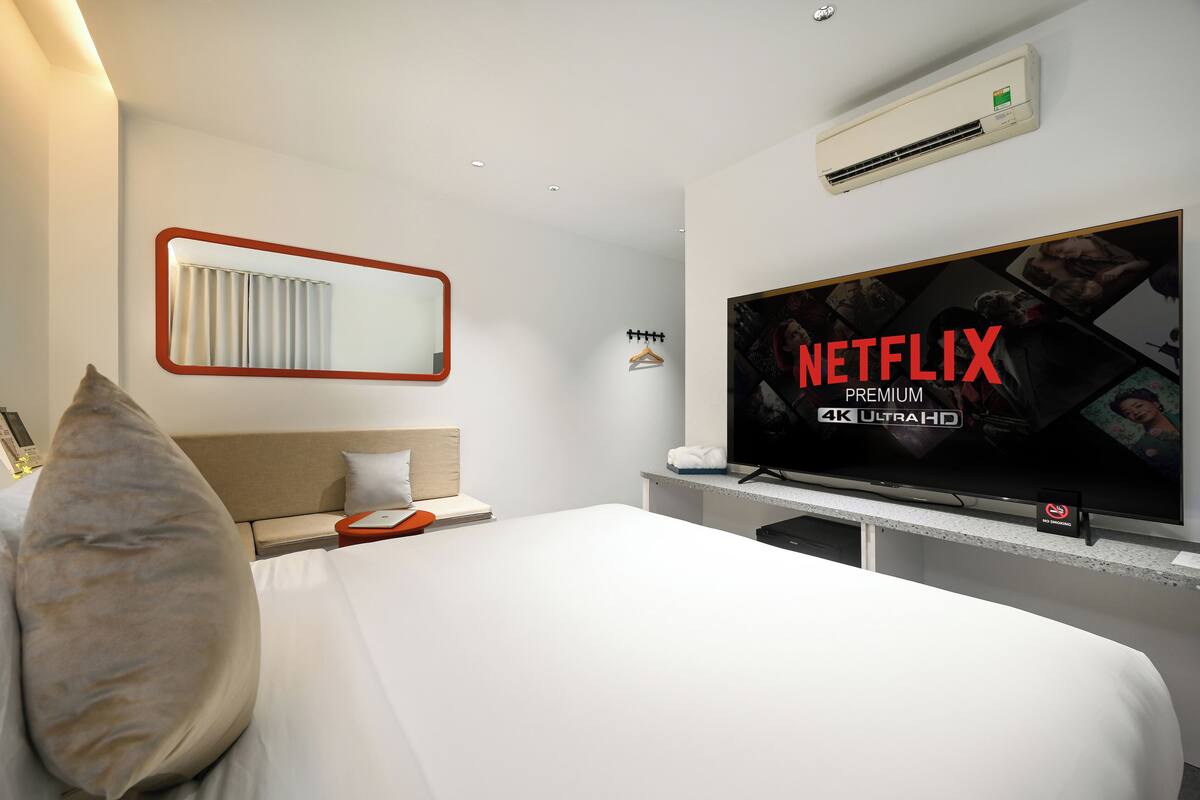 Quarto de hotel com uma televisão de tela plana grande exibindo 'NETFLIX PREMIUM 4K ULTRAHD', uma cama com lençóis brancos, uma cabeceira, uma mesinha de cabeceira com abajur e um aparelho de ar condicionado na parede.