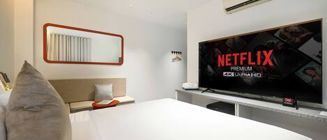 A hotel room with a large flat-screen TV displaying 'NETFLIX PREMIUM 4K ULTRAHD', a bed with white linens, a headboard, a bedside table with a lamp, and an air conditioning unit on the wall.