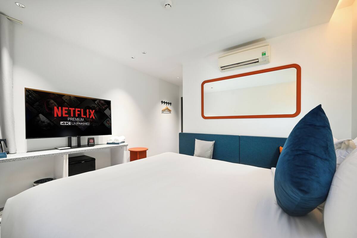 Quarto de hotel moderno com uma cama grande, uma TV de tela plana exibindo 'NETFLIX PREMIUM 4K ULTRA HD', um ar-condicionado fixado na parede e uma mesa de cabeceira com uma luminária.