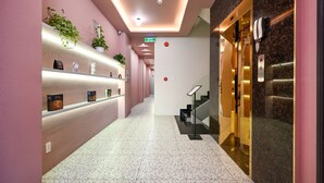 Un couloir d’hôtel au design moderne, avec un long espace de réception comprenant un comptoir, des plantes en pot et un escalier menant à un ascenseur.