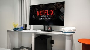 Une chambre d’hôtel moderne équipée d’un grand téléviseur à écran plat diffusant une publicité Netflix, d’un petit bureau avec une machine à café, d’une poubelle et d’un mini-réfrigérateur.