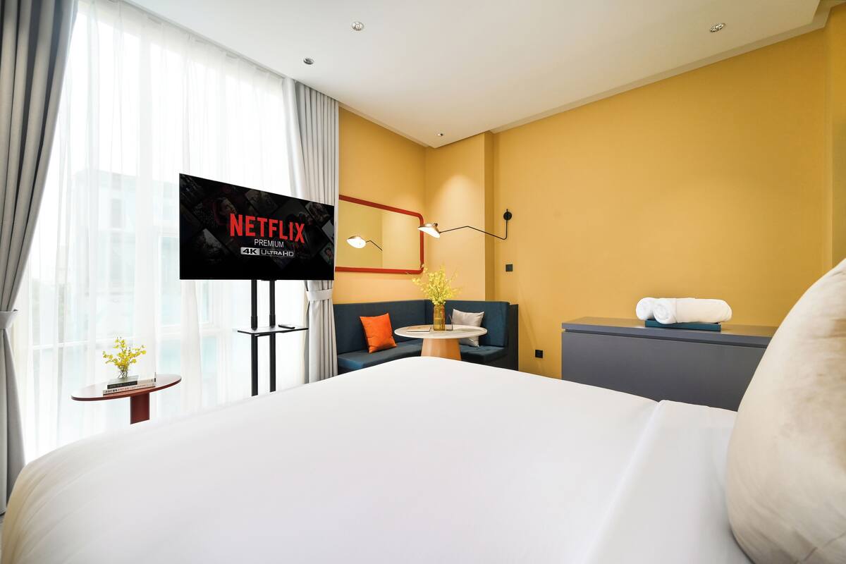 Quarto de hotel com uma cama grande, televisão em um suporte, sofá, mesinha com um vaso e espelho fixado na parede.