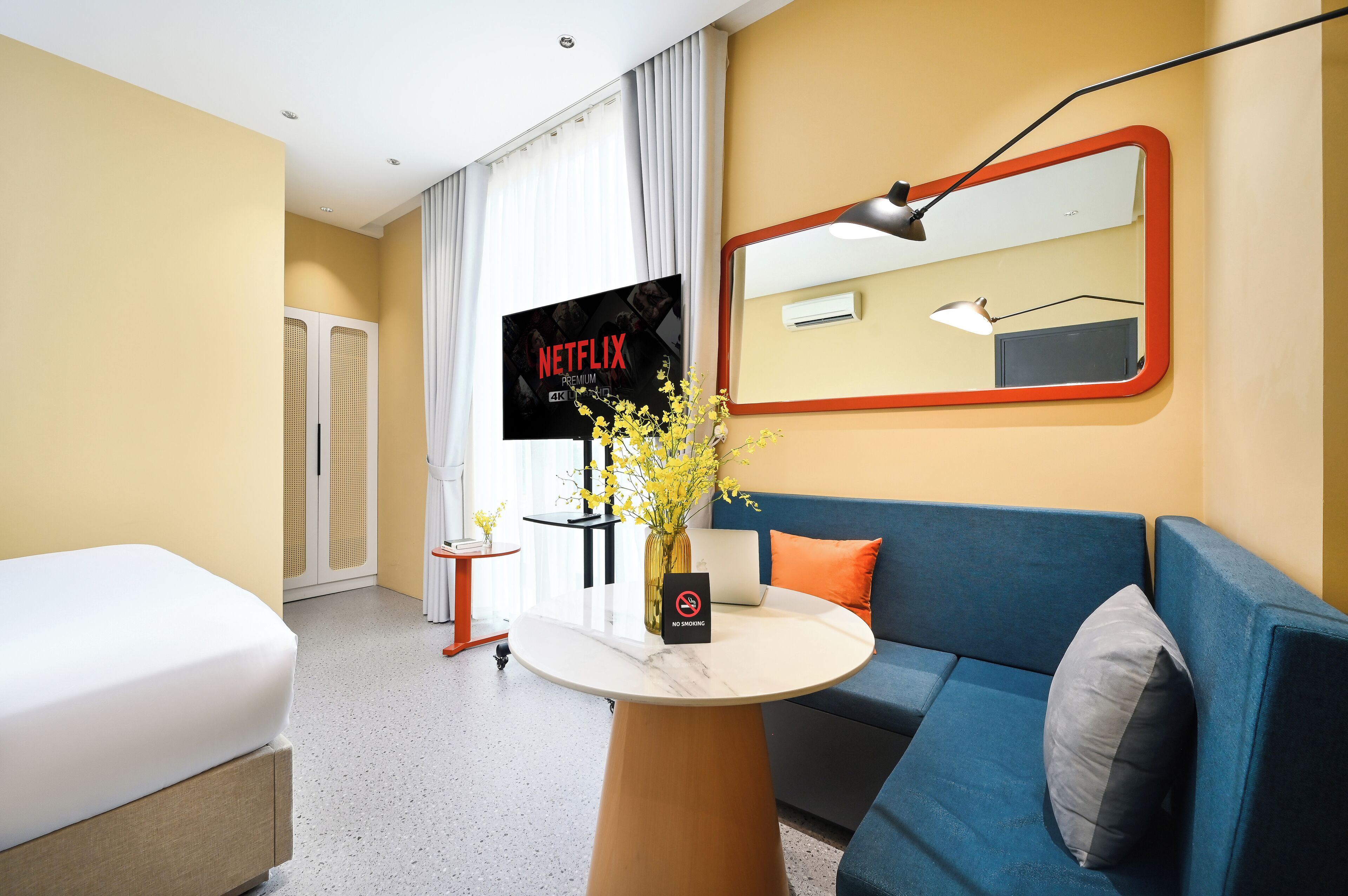 Een hotelkamer met een bed, een blauwe bank, een tafeltje met een bloevas, een televisie waarop een Netflix-advertentie wordt vertoond, en een spiegel met een rode lijst.
