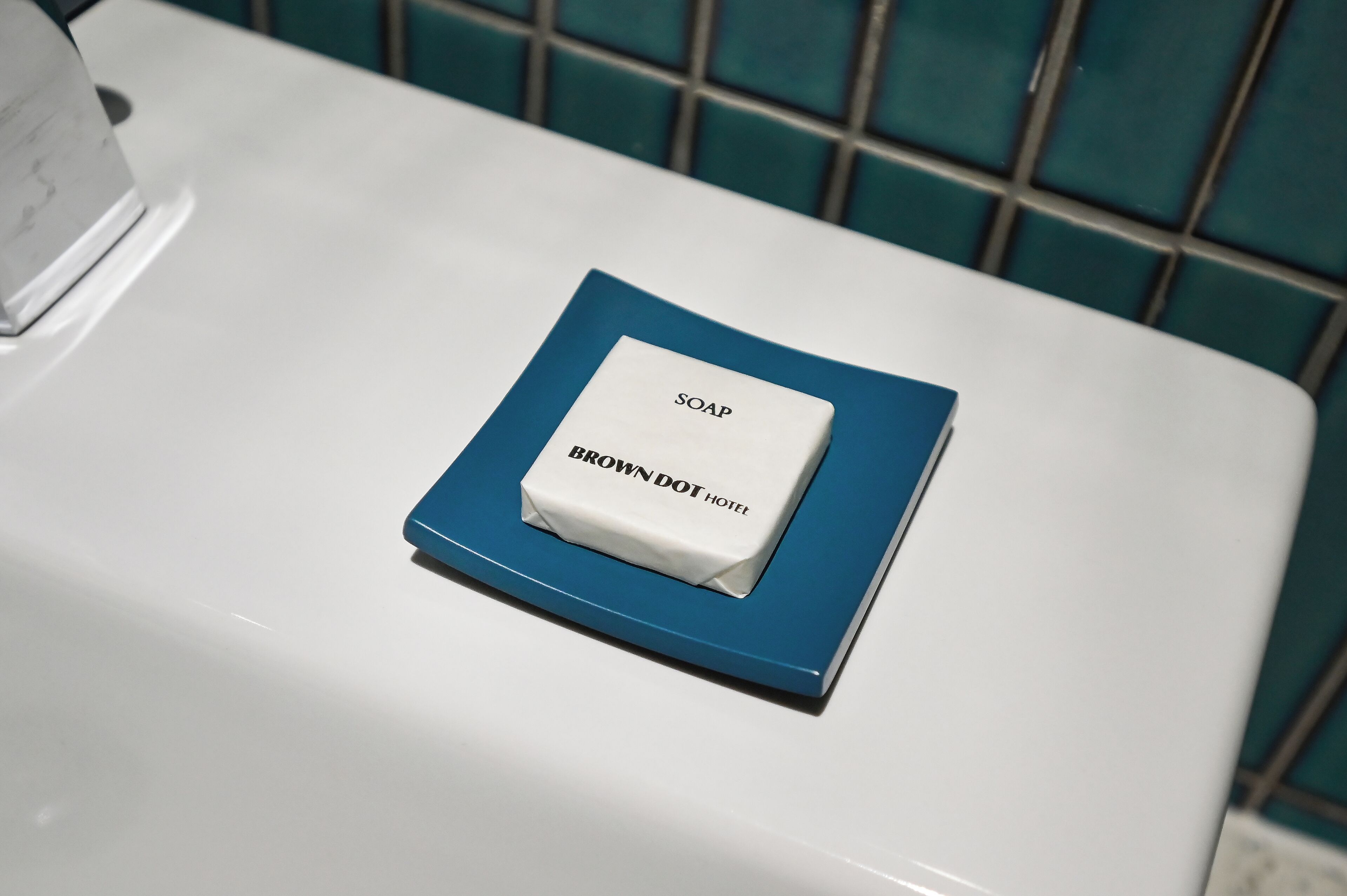 Un lavamanos blanco con un plato para jabón azul que contiene una barra de jabón con la etiqueta 'SOAP BROWN DOT HOTEL'.