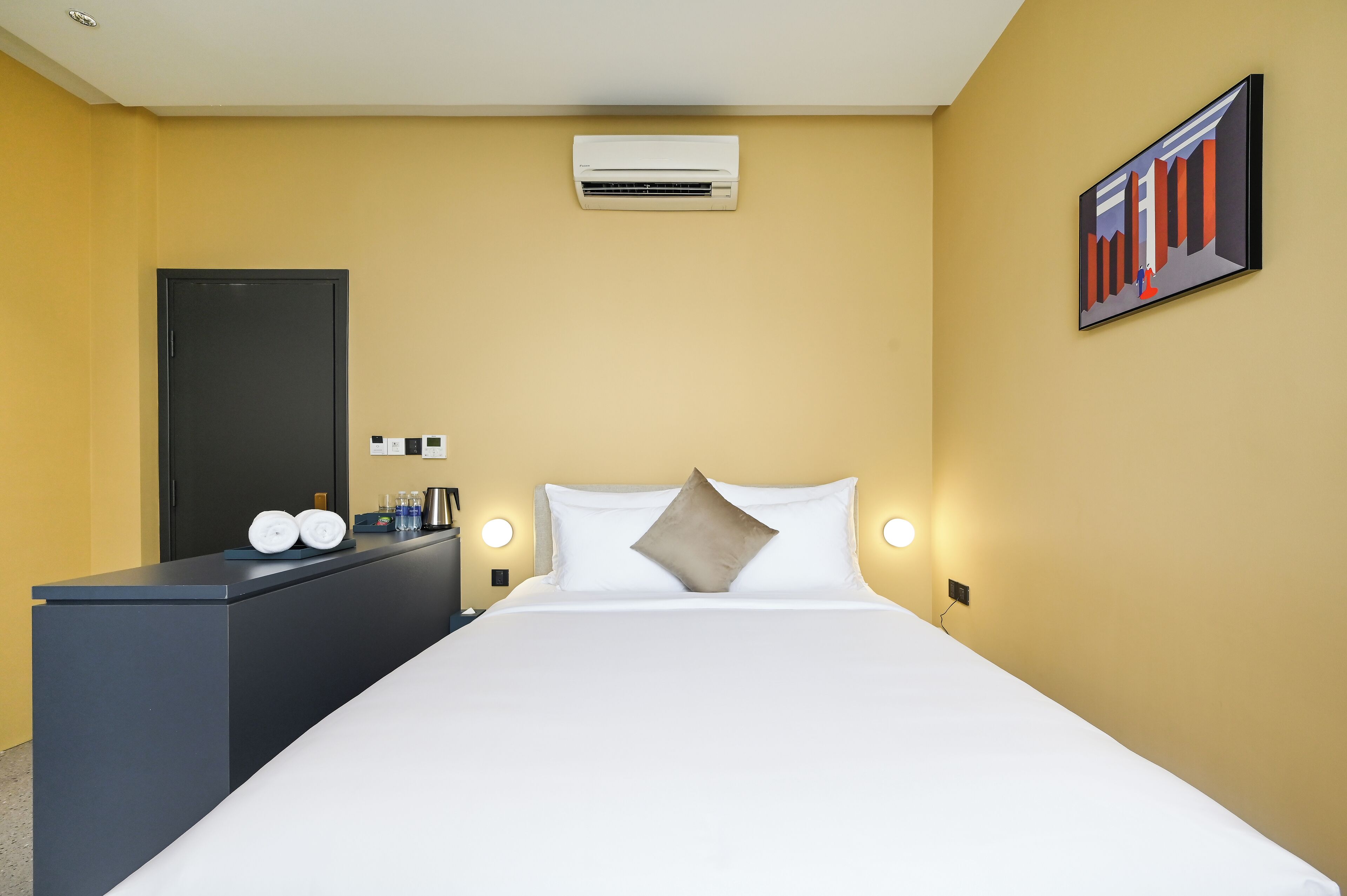 A hotel room with a bed, a desk with a chair, a wall-mounted air conditioner, a painting, and a door.