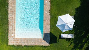 Luftaufnahme eines Schwimmbeckens mit Steinrand, umgeben von Rasen, sowie einem weißen Sonnenschirm und einem Stuhl in der Nähe.