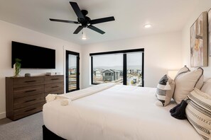 A bedroom with a bed, a ceiling fan, a TV, a dresser, and a large window with a view.