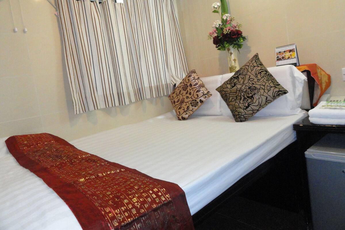 Quarto de hotel com uma cama de solteiro, cortinas listradas, um arranjo floral e uma pequena escrivaninha com cadeira.