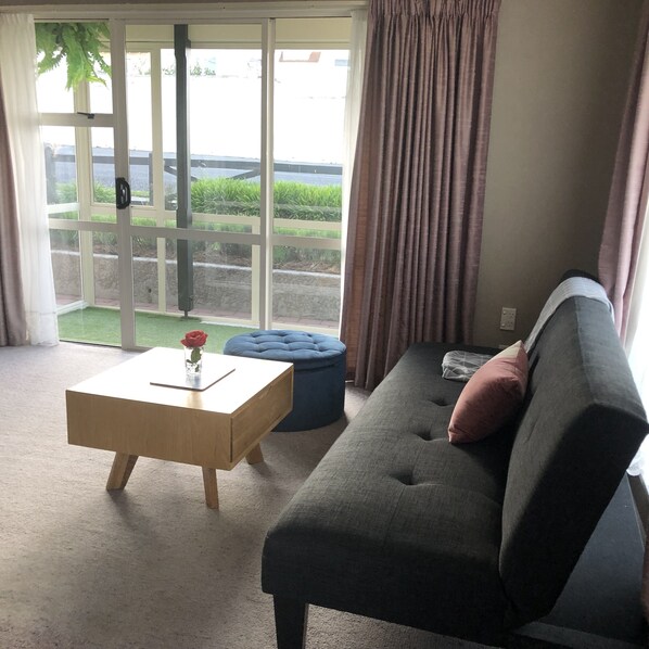 A living room with a grey sofa, a wooden coffee table with a rose vase, and a large sliding glass door leading to a garden.