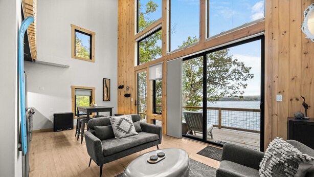 A modern living room with a large window overlooking a lake, a gray sofa, a coffee table, and a wooden deck outside. - Black Diamond: "Waterfront Finger Lakes Home with Hot Tub!" (Hector)
