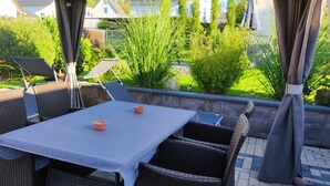 En udendørs terrasse med et bord og stole, omgivet af grønt og udsigt til huse.