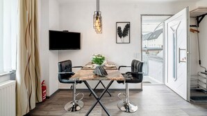 Et spiseområde med et træbord, to stole, en hængende lampe, et fladskærms-tv og et billede af et efterårblad i ramme.