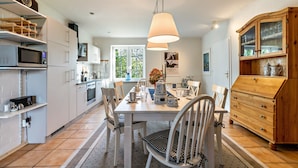 Una cocina moderna con armarios blancos, una mesa de comedor con sillas, un mueble de madera con puertas de vidrio y una lámpara colgante.
