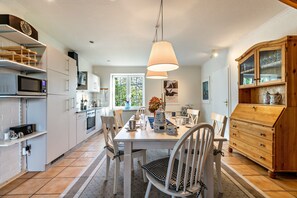 A modern kitchen with white cabinetry, a dining table with chairs, a wooden cabinet with glass doors, and a hanging light fixture. - Hygge Frisian House by the Sea with Private Garden and Terrace (Nordstrand)