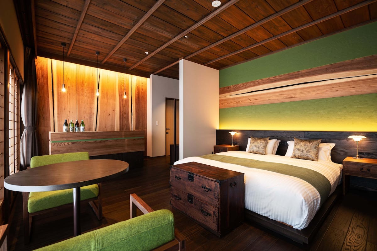 A modern hotel room with a wooden ceiling, a round dining table, green upholstered chairs, a large bed with multiple pillows, and a nightstand with a lamp.