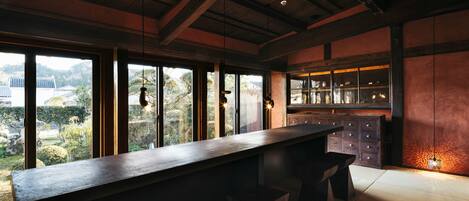 A modern kitchen with a long counter, wooden cabinets, and large windows offering a view of the surrounding landscape.