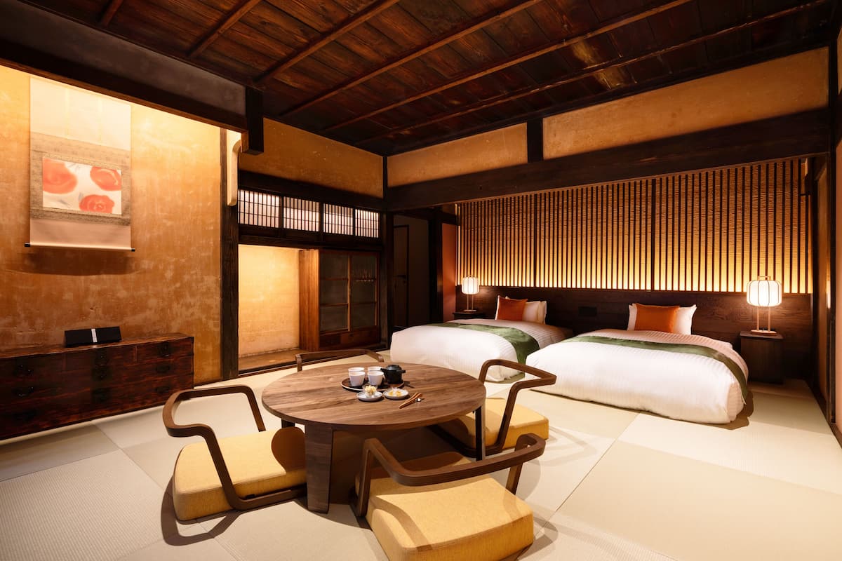 A traditional Japanese-style room with tatami flooring, a low wooden ceiling, and a bed with a green and orange blanket. There is a round table with a tea set and a wooden cabinet with drawers.