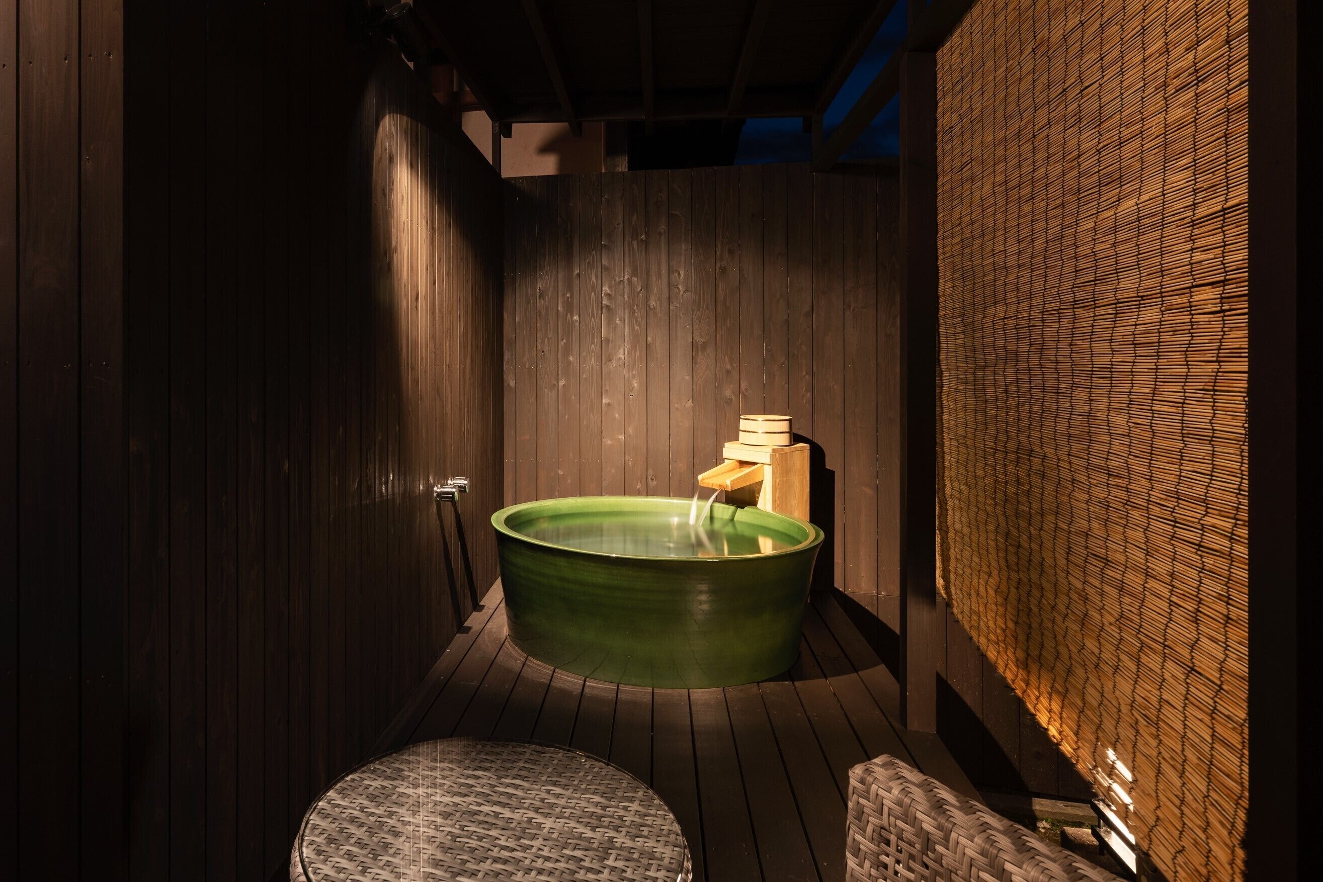 A wooden-floored spa room with a green hot tub, a small wooden bench, and a woven wall.