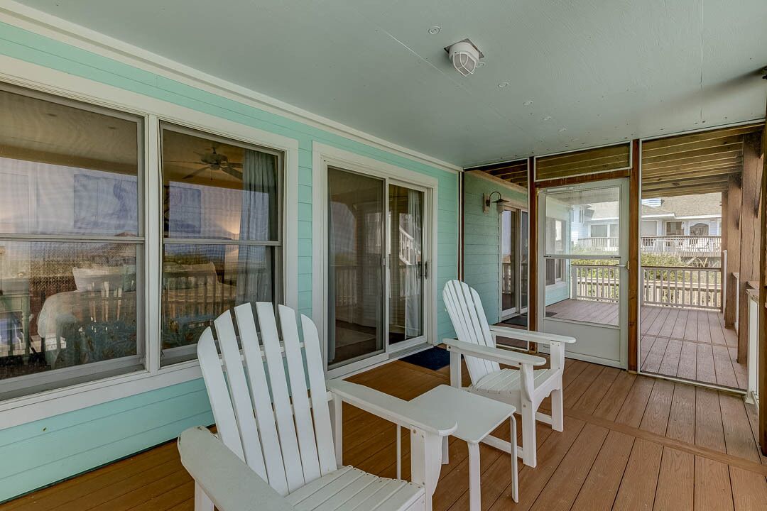 A covered porch with white Adirondack chairs and a small table, large sliding glass doors leading to a deck, and a ceiling fan.